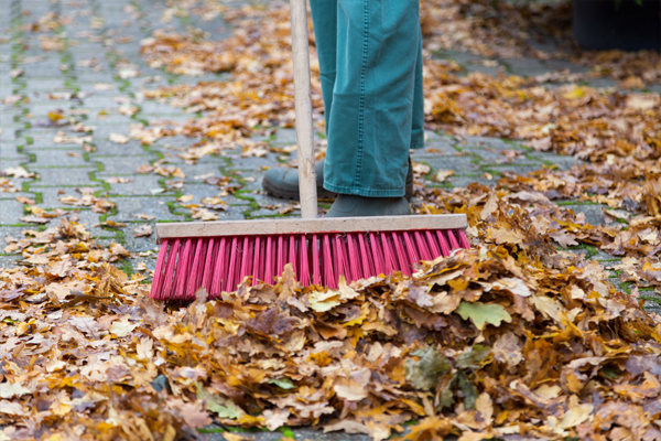Ein Besen fegt buntes Herbstlaub auf einer gepflasterten Hoffläche zusammen.
