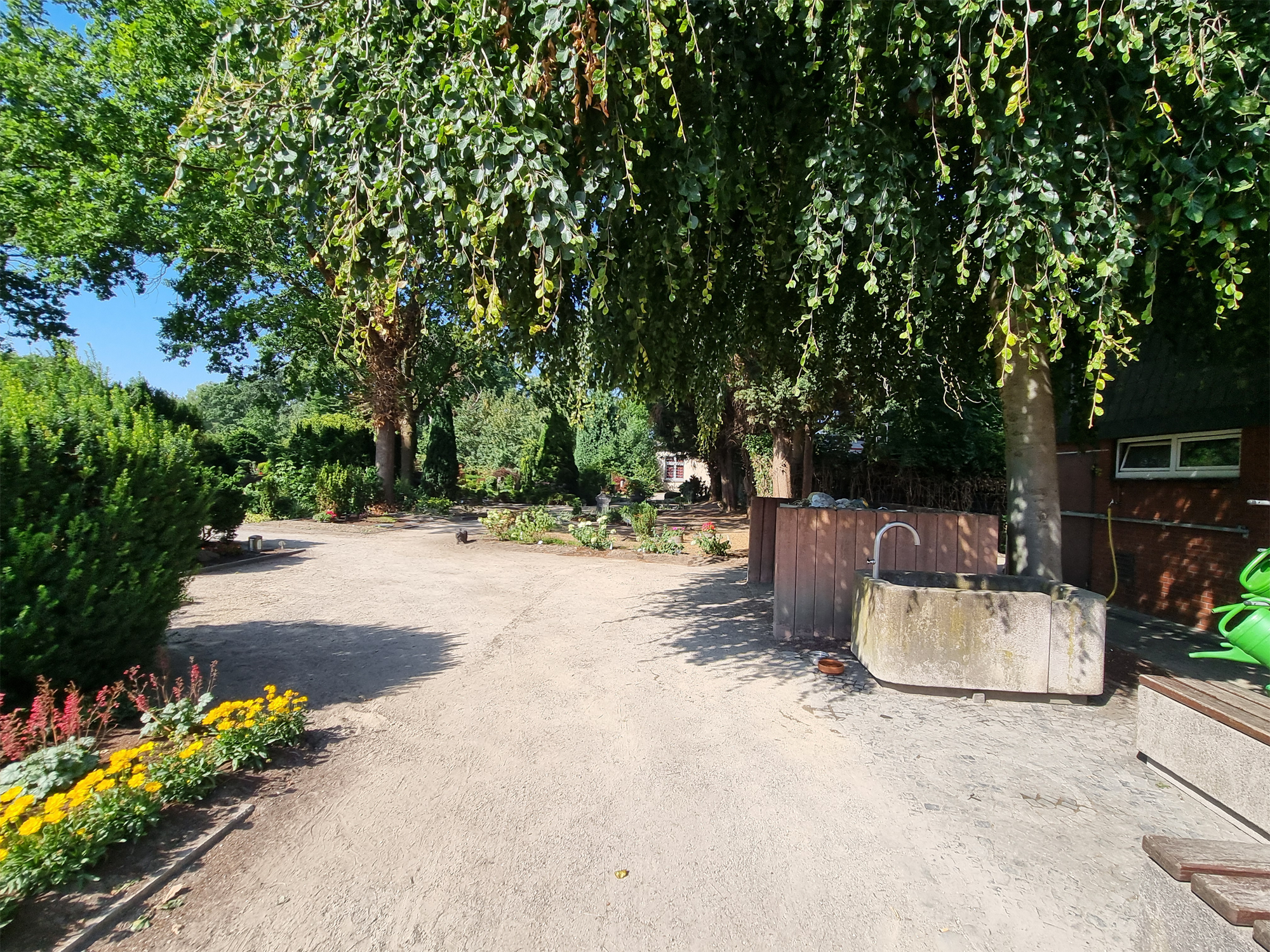Brunnen vor alten, schattigen Bäumen und Beeten mit bunt blühenden Blumen.