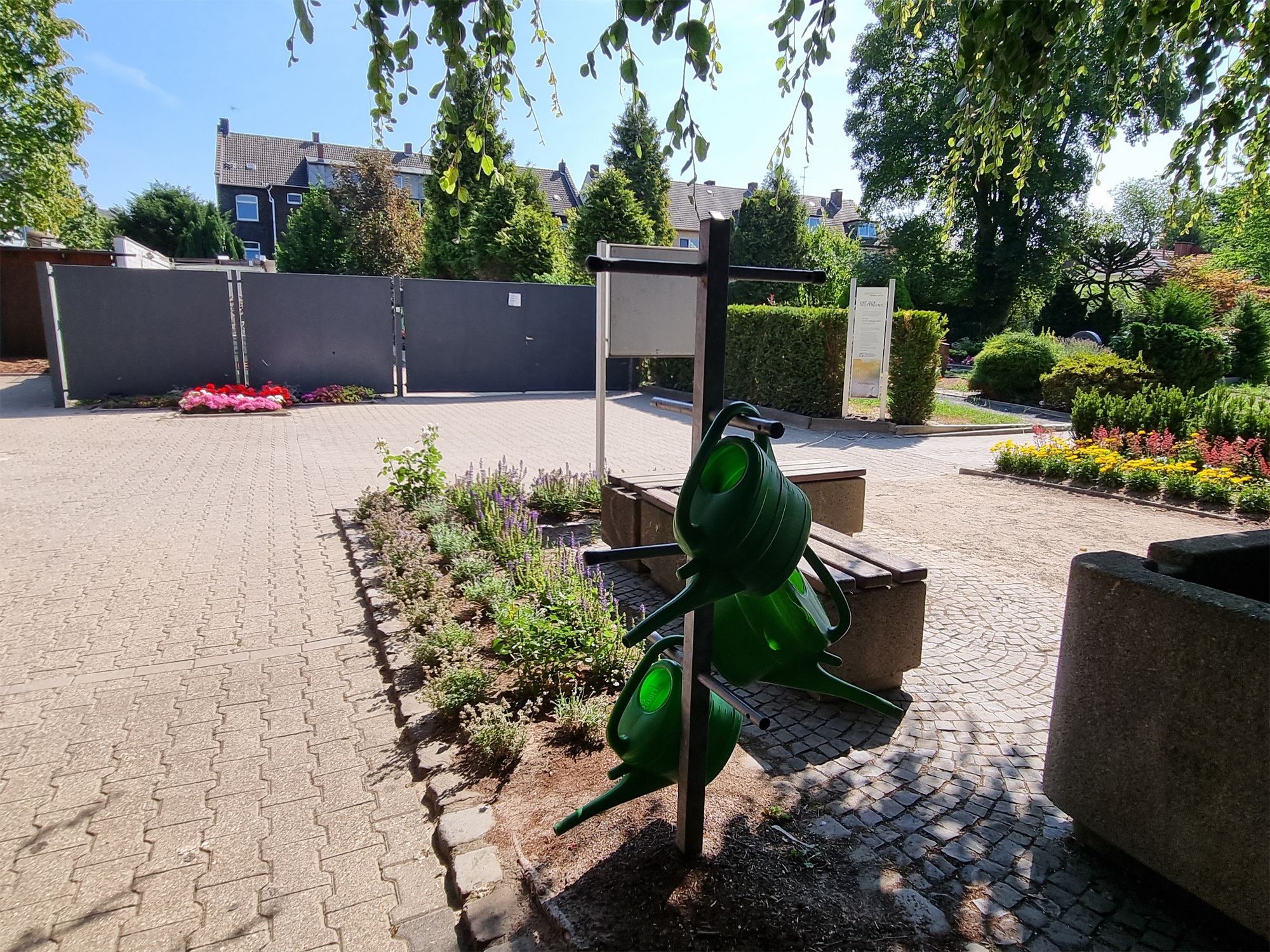 Im Vordergrund Brunnen mit Gießkannen, dahinter Beete mit bunten Blumen vor dem Betriebshof.