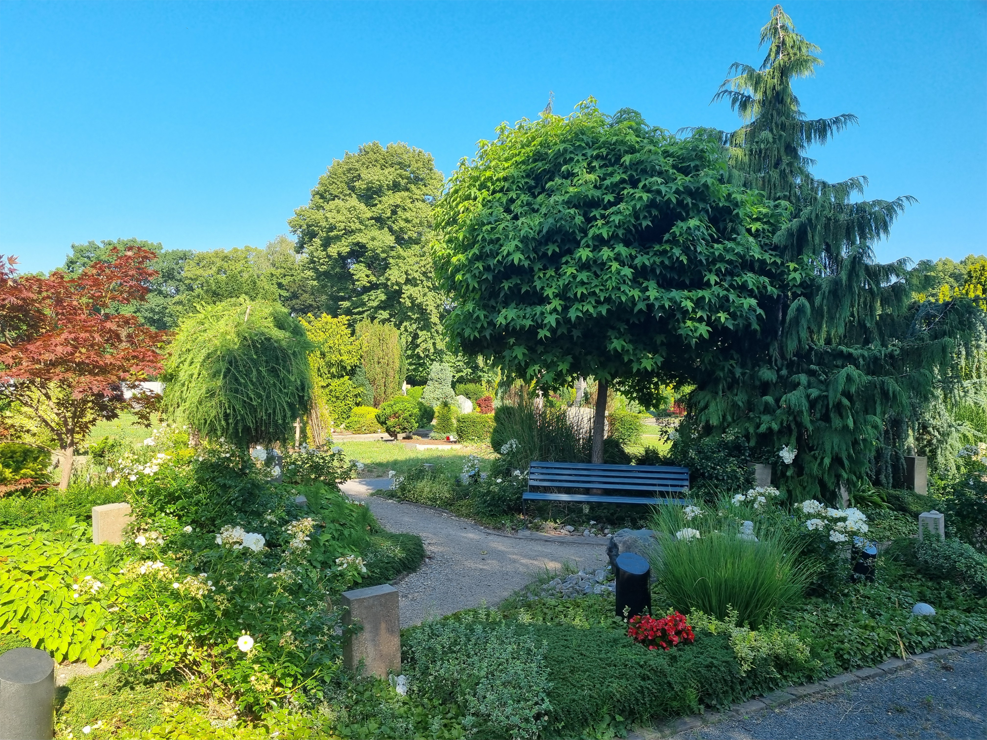 Parkähnlich gestaltete Grabanlage mit Grabsteinen, bunten Blumen und einer Bank vor grünen Bäumen.