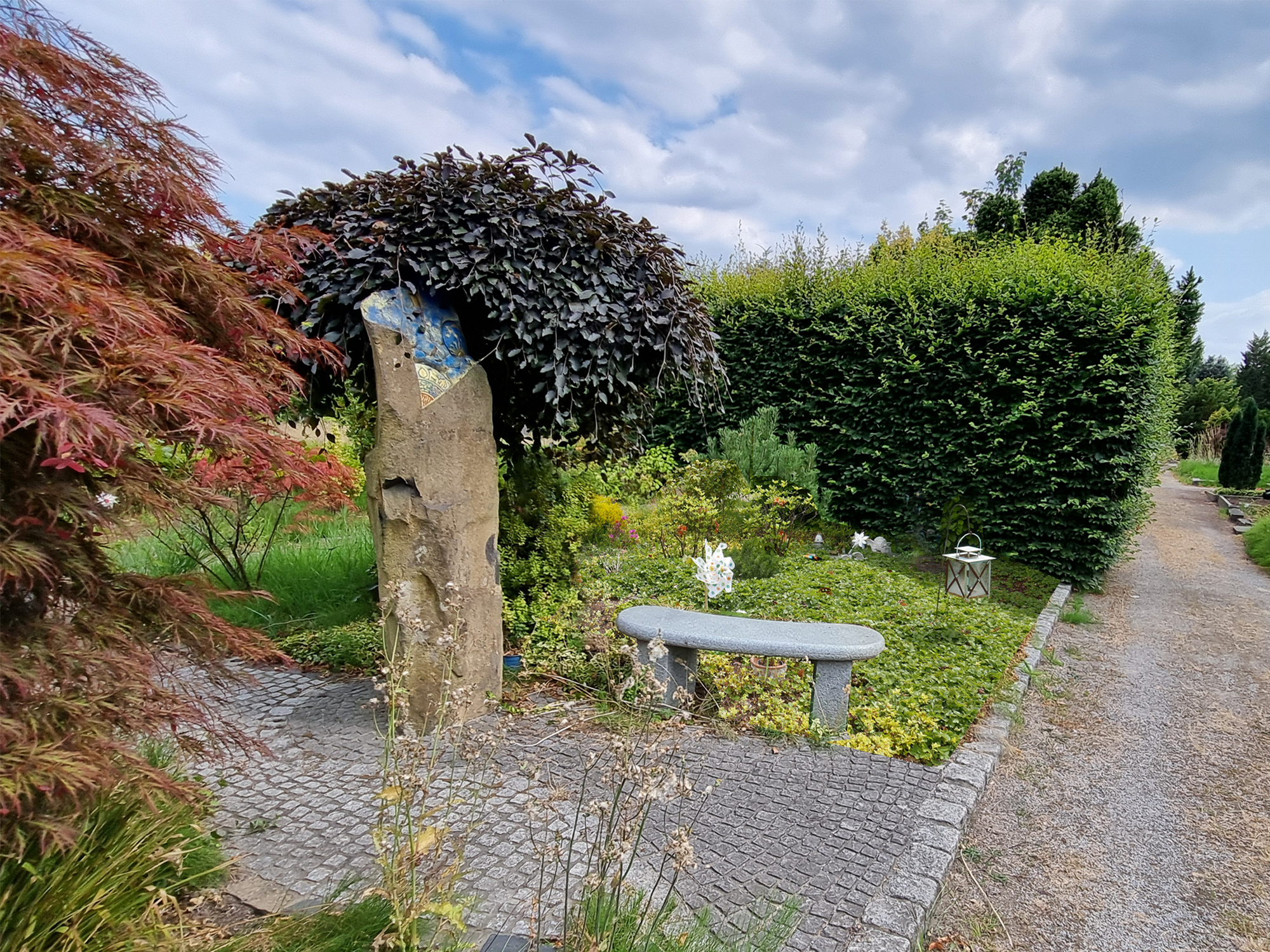 Kindergrabfeld mit Bank und Skulptur vor blühenden Pflanzen und einer grünen Hecke.