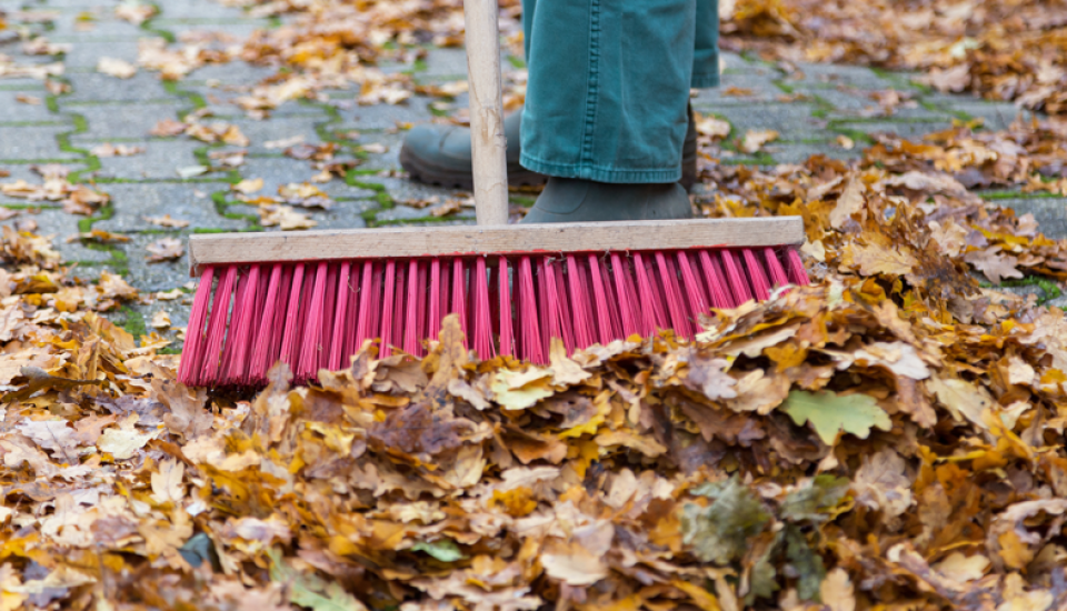 Ein Besen fegt buntes Herbstlaub auf einer gepflasterten Hoffläche zusammen.