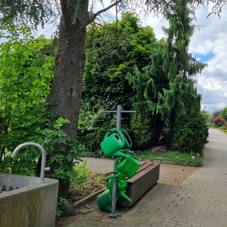 Friedhofsbrunnen mit Gießkannenständer und Sitzbank.